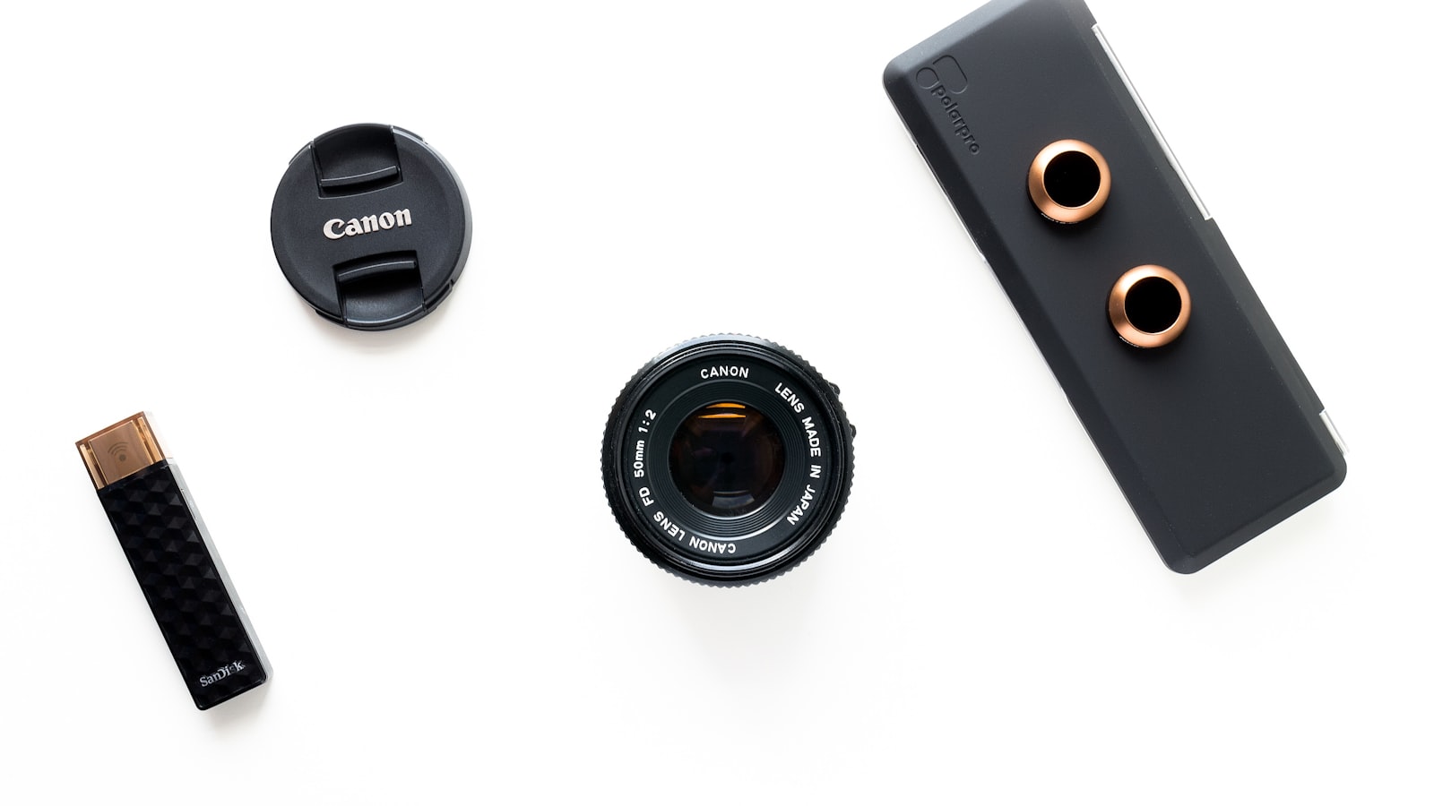 Camera lens, lens cap, USB drive and a polariser case laid out flat on a clean white surface — a product-photography flatlay.